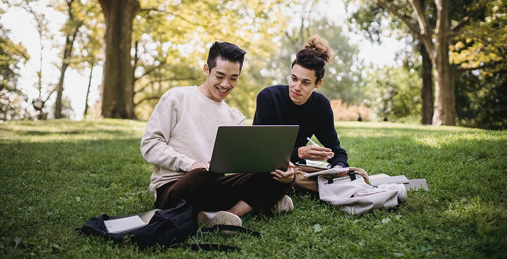 two college students sitting on green grass field using laptop computer