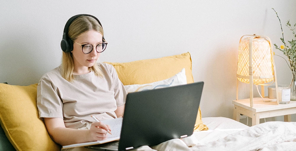 female remote employee working on laptop from her bed wearing headphones