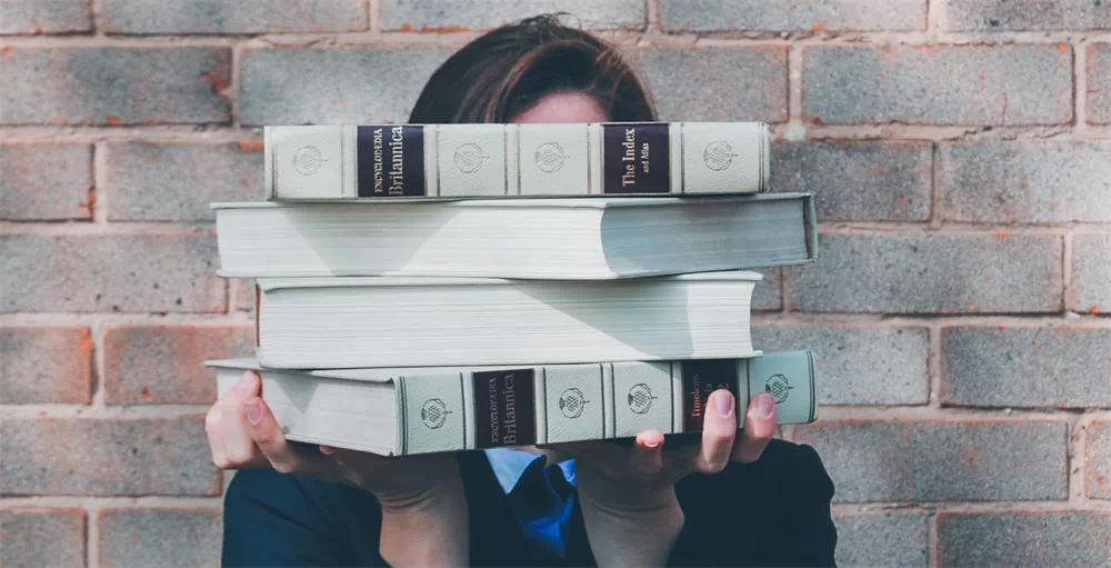 college student holding pile of financial education books covering face